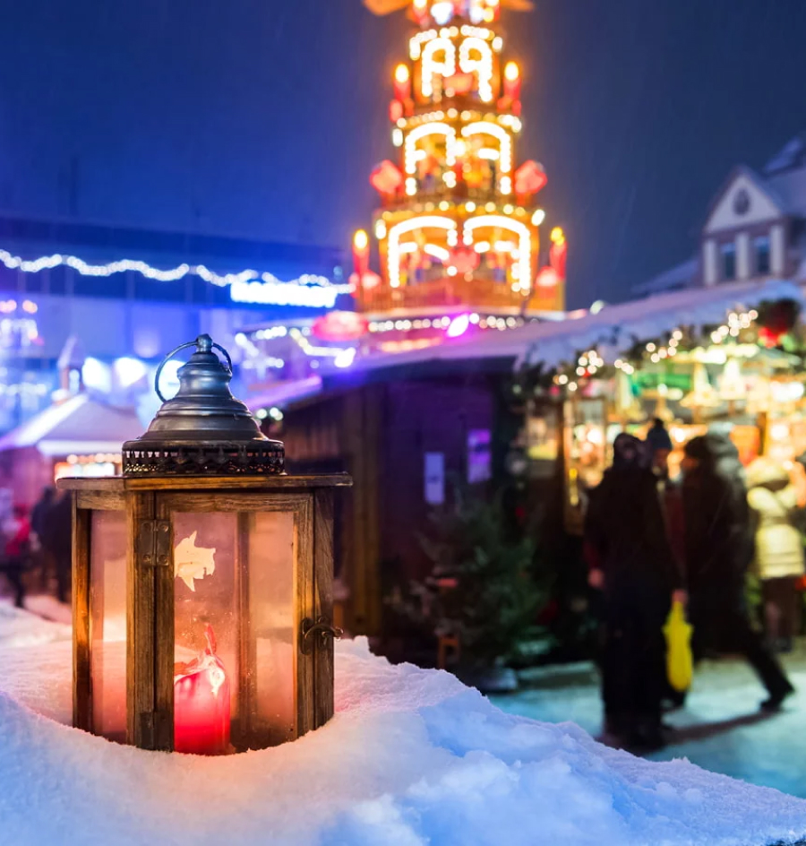 Eine Kerze steht im Schnee vor einer beleuchteten Weihnachtsmarktkulisse
